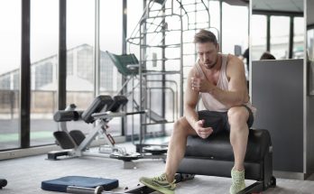 Tired young muscular male athlete in casual sportswear and sneakers sitting on bench and resting after intense weightlifting