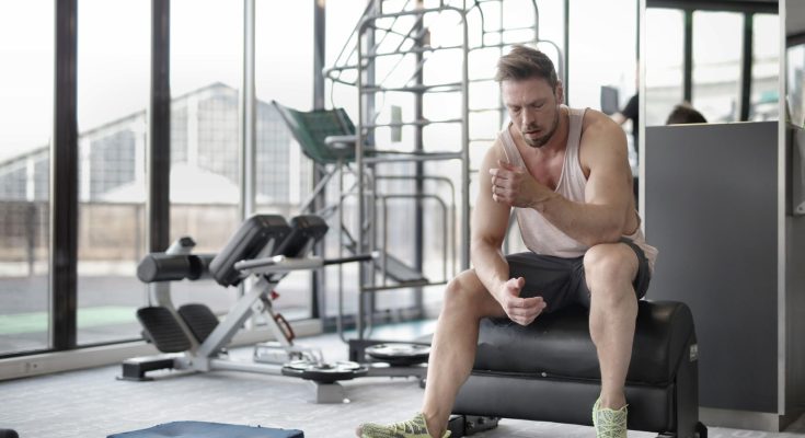Tired young muscular male athlete in casual sportswear and sneakers sitting on bench and resting after intense weightlifting