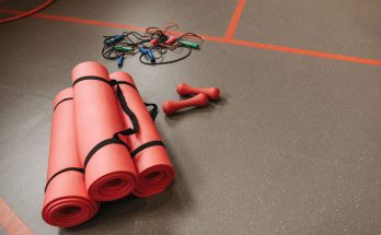 Red yoga mats, jump ropes, and dumbbells neatly arranged on a gym floor for fitness activities. — Fitness Weight Training - W