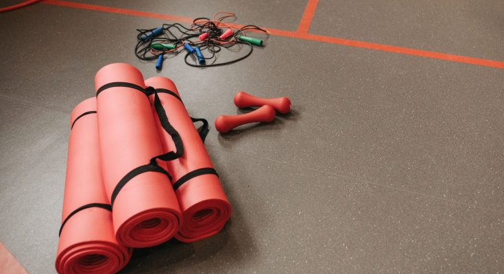 Red yoga mats, jump ropes, and dumbbells neatly arranged on a gym floor for fitness activities. — Fitness Weight Training - W