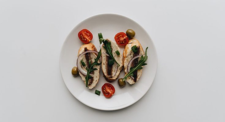 A top view of delicious bruschetta with anchovies, olives, and cherry tomatoes on a white plate. — Puppy Food - Setting It Up